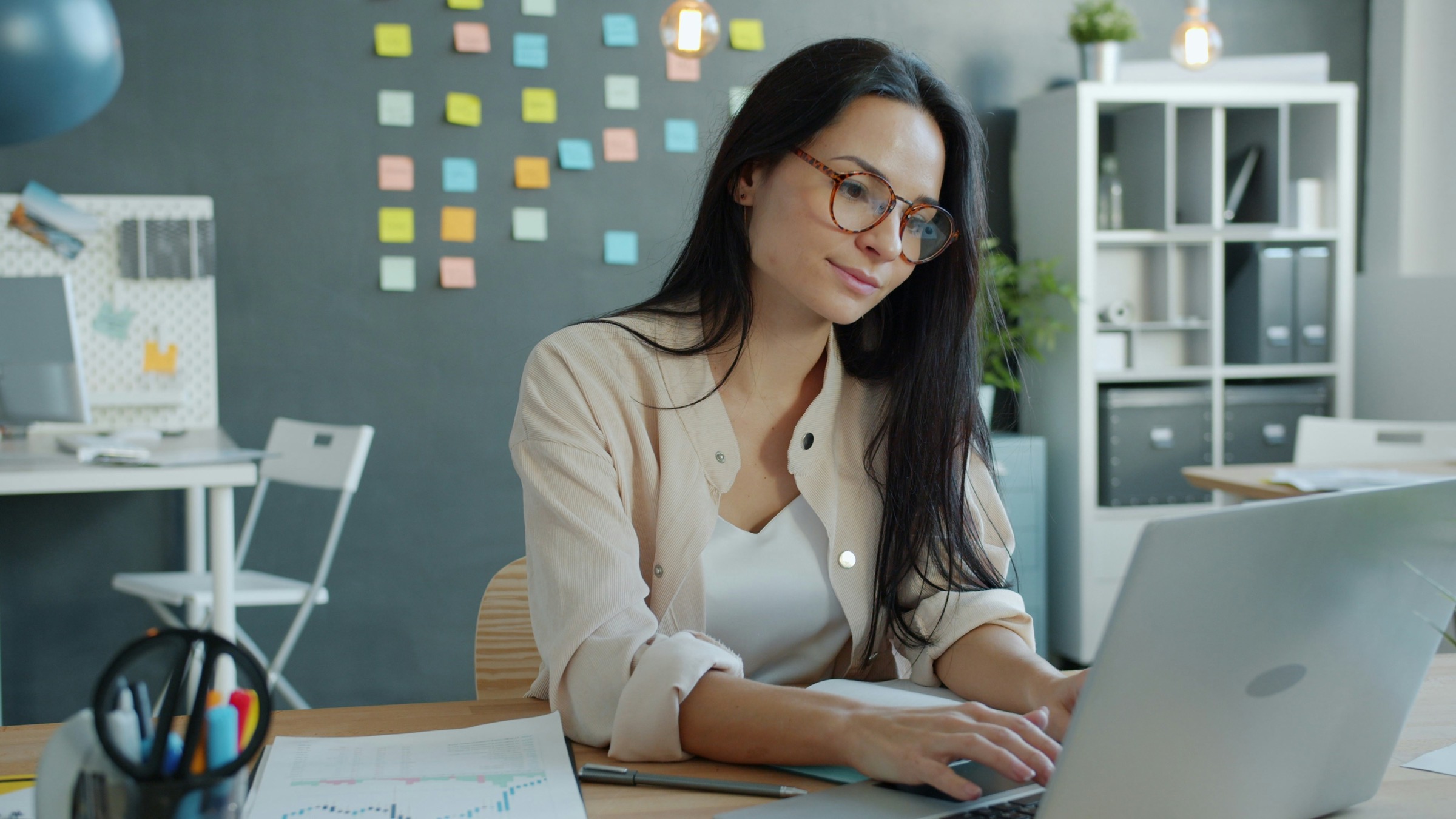 A learning designer at her desk, working on her laptop, sticky notes on the wall behind her.